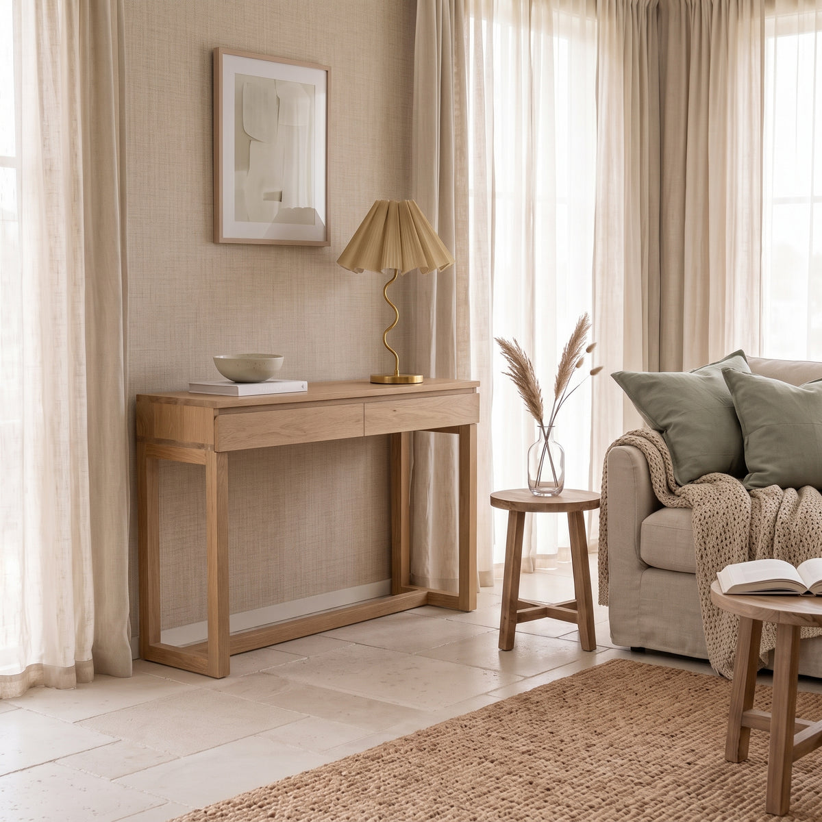 A light natural wood console table in a styled living room setting, featuring a pleated beige table lamp, a small ceramic bowl, and a stack of books on top.