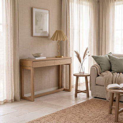 A light natural wood console table in a styled living room setting, featuring a pleated beige table lamp, a small ceramic bowl, and a stack of books on top.