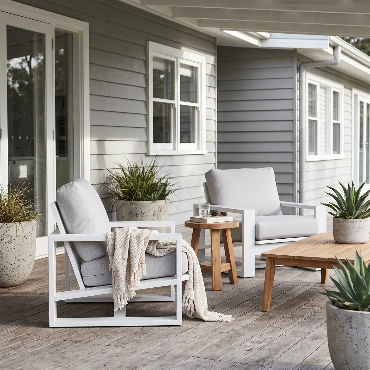 A styled outdoor patio seating area featuring two white-framed lounge chairs with light gray cushions, a small round wooden side table, a wooden coffee table, and potted plants against a gray house siding background.