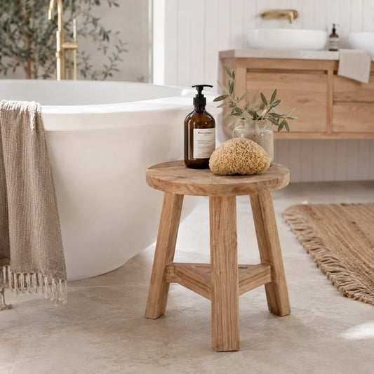 A small round wooden stool in a bright, modern bathroom setting next to a white freestanding bathtub. The stool is styled with an amber glass bottle, a natural sponge, and a small glass vase with greenery.