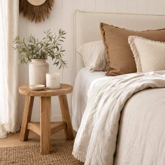 A styled bedroom scene showing a bed with cream bedding, a tan textured pillow, and a white fringed pillow, next to a small wooden stool holding a white vase with greenery and a candle.
