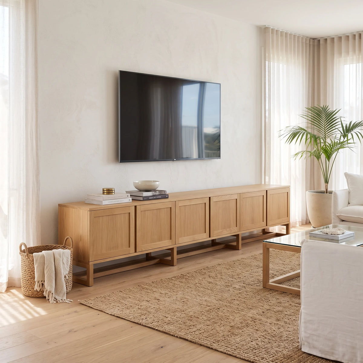 A long, low light-wood media console with six paneled cabinet doors stands against a cream wall in a bright living room. A black flat-screen TV is mounted above it, with books and small decor objects placed on top.