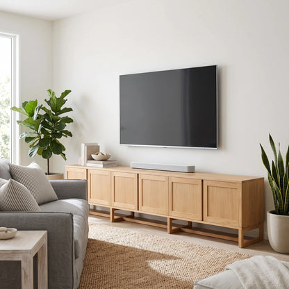 A light natural wood TV stand with multiple cabinet doors and open lower shelves, styled in a modern living room with a gray sofa, potted plants, and a wall-mounted flat-screen TV.