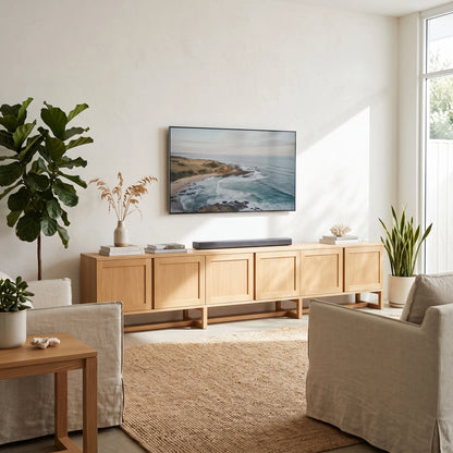 A modern living room scene with a light wood media console beneath a wall-mounted TV showing a coastal landscape. The room includes upholstered chairs, potted plants, a side table, and a woven rug in a bright, sunlit interior.