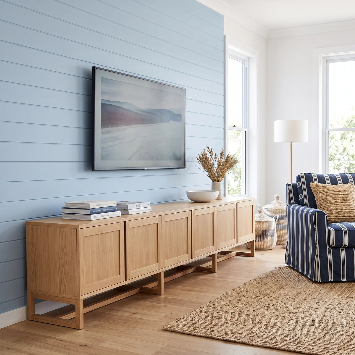 A long light oak wood sideboard console with six cabinet doors is styled in a coastal living room against a light blue shiplap wall, with a framed landscape print mounted above it.