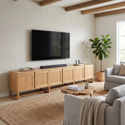 A light natural wood media console with five cabinet doors stands against a white brick wall in a bright living room, holding a large black TV and a soundbar. A round wooden coffee table, woven rug, gray sofa, and a potted fiddle-leaf fig plant are also visible.