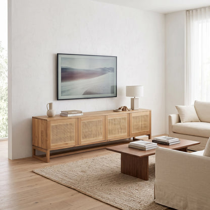 A light natural wood sideboard with woven cane panels stands in a bright, modern living room against a white wall, displaying a framed landscape print. The room is styled with a beige sofa, a wooden coffee table, a textured rug, and a table lamp.