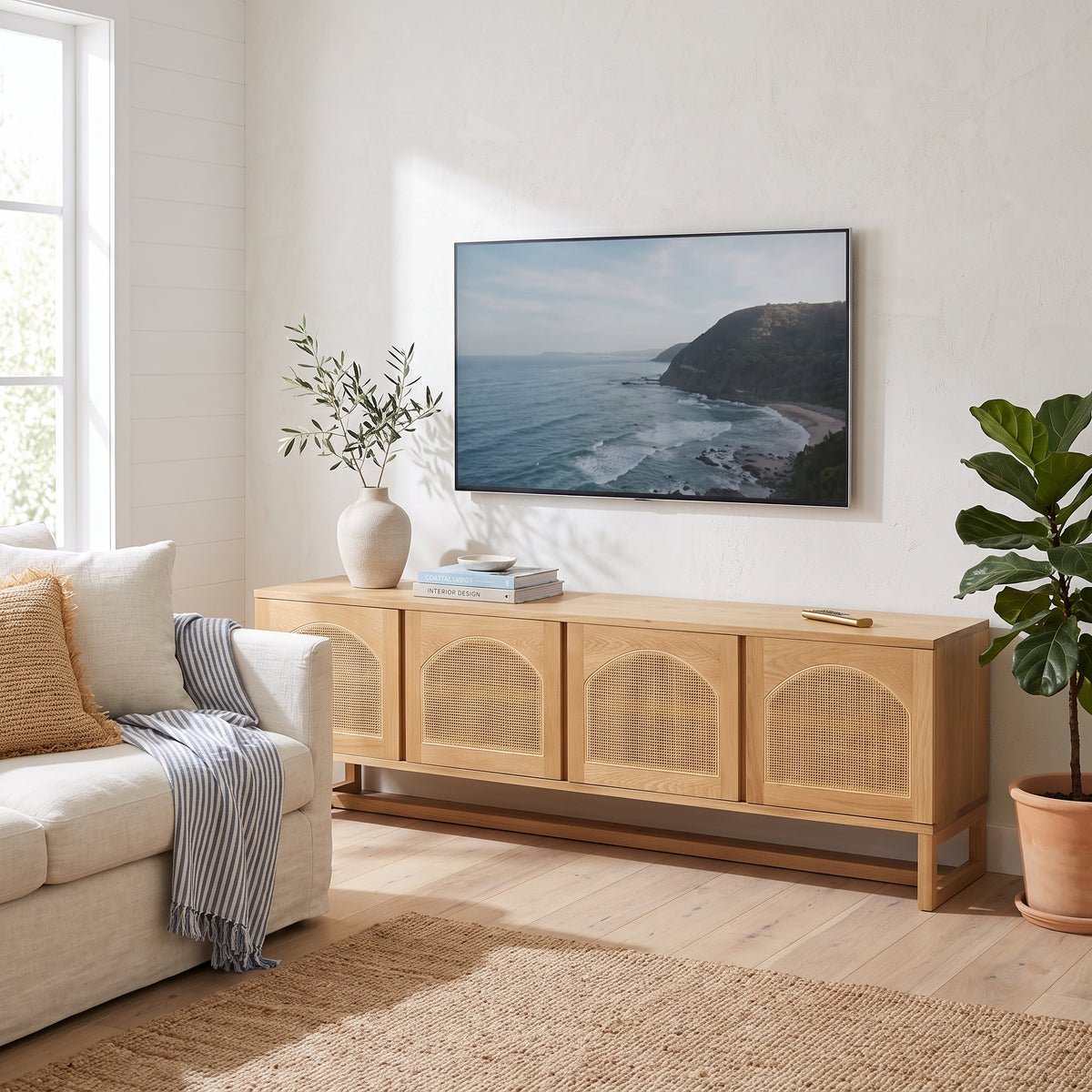 A light natural wood TV stand or media console with three arched cane-screen doors, styled in a bright living room with a sofa, a large framed coastal landscape print, and a potted plant.