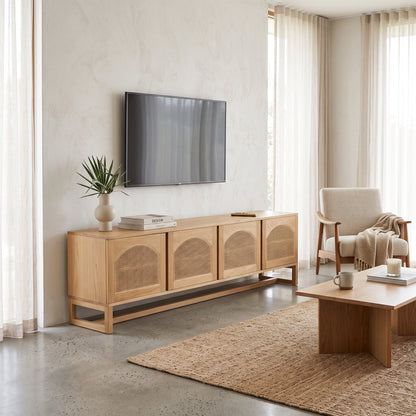 A light natural wood TV console with woven cane doors stands in a bright, modern living room against a textured beige wall, with a wall-mounted TV above it.