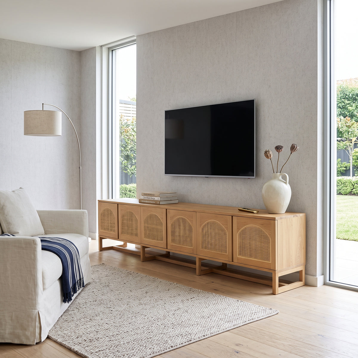 A light natural wood media console with woven cane doors, styled in a bright, modern living room with a wall-mounted flat-screen TV, a beige armchair, and a large textured rug.