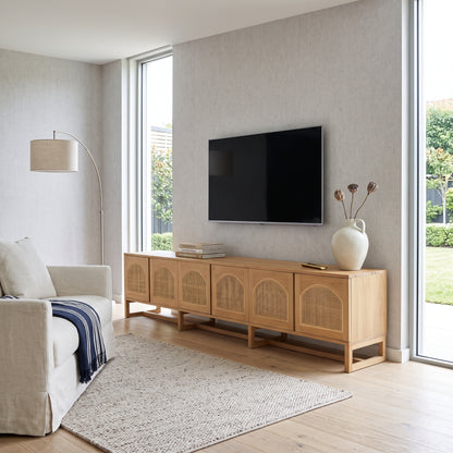 A light natural wood media console with woven cane doors, styled in a bright, modern living room with a wall-mounted flat-screen TV, a beige armchair, and a large textured rug.