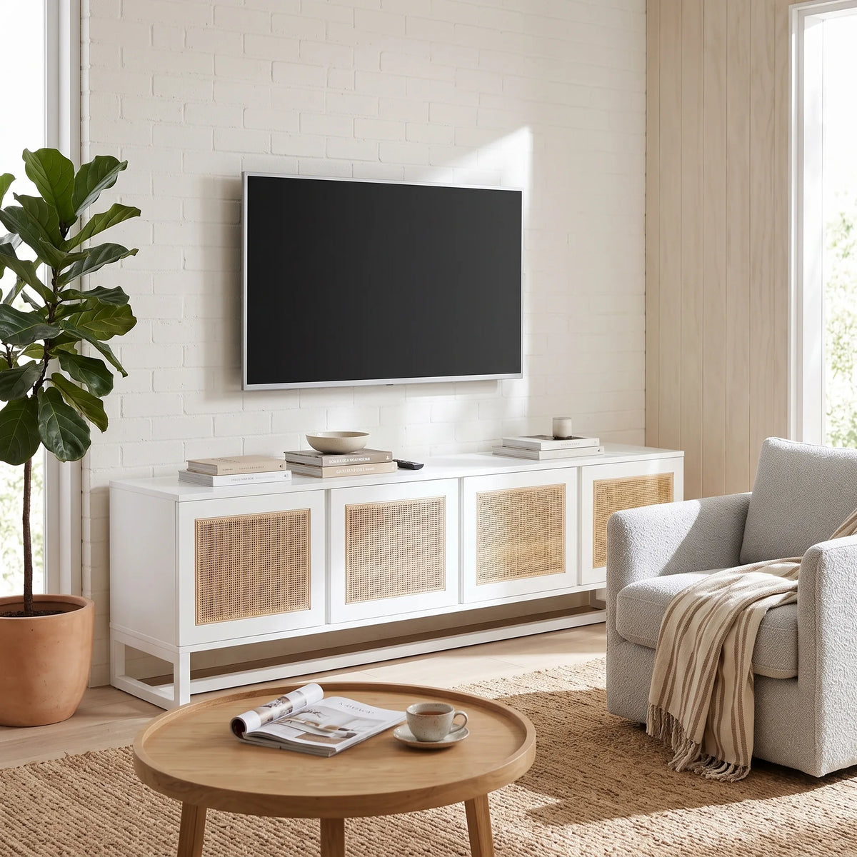 A modern white TV stand with woven cane panels in a bright living room, featuring a wall-mounted TV, a round wooden coffee table, a light gray armchair, and a potted fiddle-leaf fig plant.