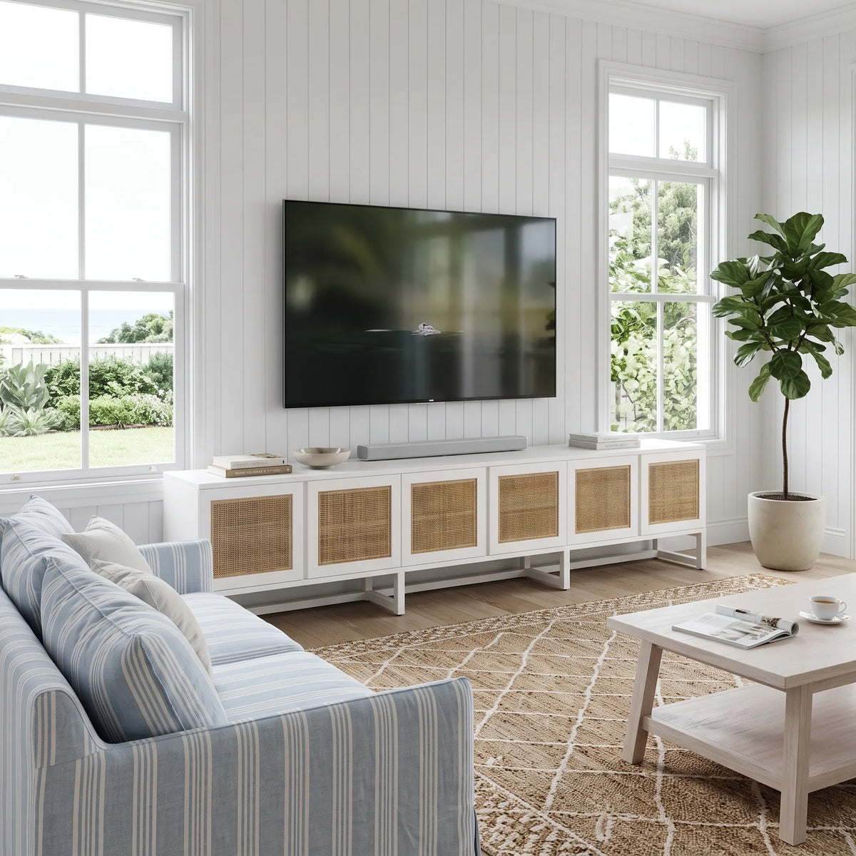A modern living room scene featuring a white media console with woven cane doors, a wall-mounted flat-screen TV, a striped blue and white sofa, a light wood coffee table, and a potted fiddle-leaf fig plant.