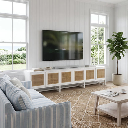 A modern living room scene featuring a white media console with woven cane doors, a wall-mounted flat-screen TV, a striped blue and white sofa, a light wood coffee table, and a potted fiddle-leaf fig plant.