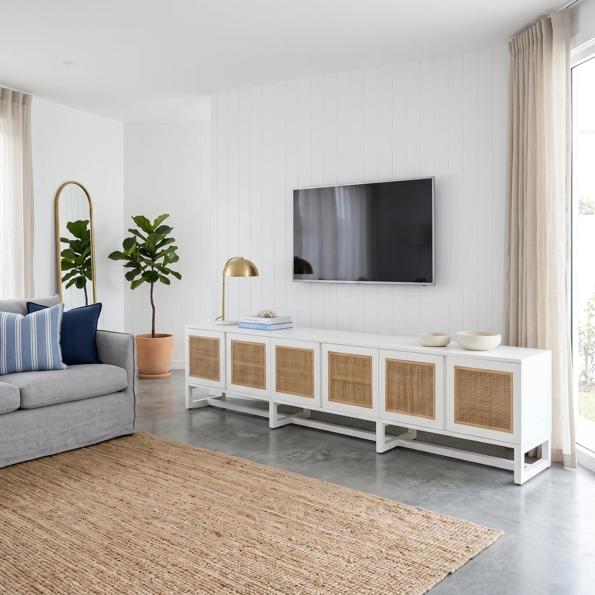 A modern white media console with woven cane doors, styled in a bright living room next to a gray sofa and a wall-mounted TV.