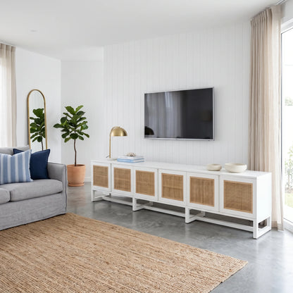 A modern white media console with woven cane doors, styled in a bright living room next to a gray sofa and a wall-mounted TV.