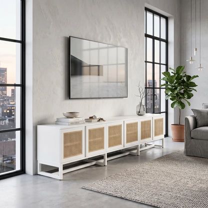 A modern white media console with woven cane doors, styled in a contemporary living room setting with a wall-mounted TV, large windows, and a potted plant.