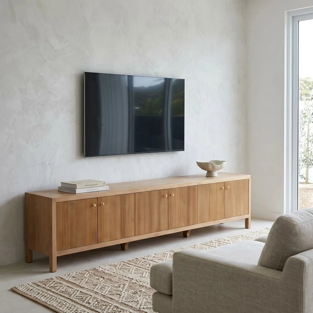 A modern light oak wood media console with vertical slat doors and small brass knobs, styled in a minimalist living room under a wall-mounted flat-screen TV.