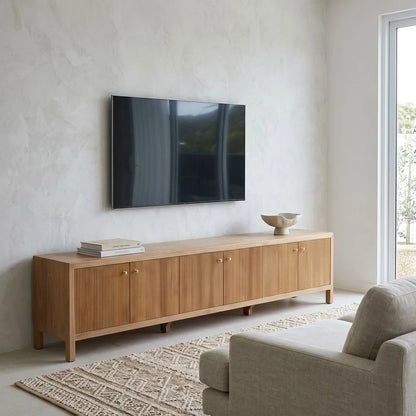 A modern light oak wood media console with vertical slat doors and small brass knobs, styled in a minimalist living room under a wall-mounted flat-screen TV.
