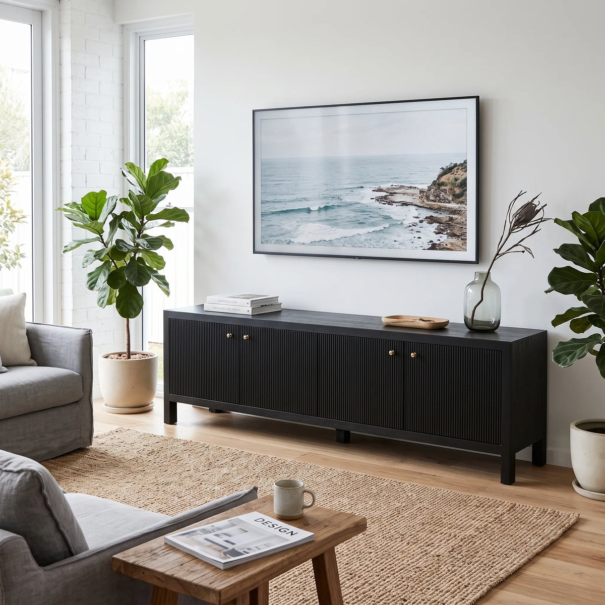 A modern black wooden media console in a bright living room, styled with a framed ocean landscape print on the wall above it, flanked by potted plants and a small wooden side table in the foreground.