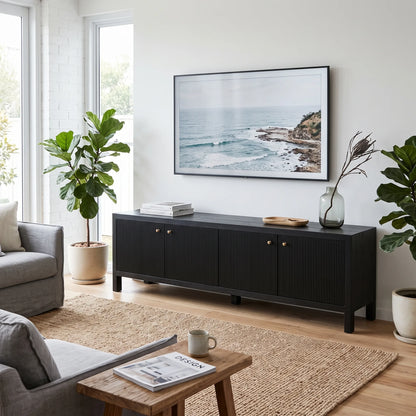 A modern black wooden media console in a bright living room, styled with a framed ocean landscape print on the wall above it, flanked by potted plants and a small wooden side table in the foreground.