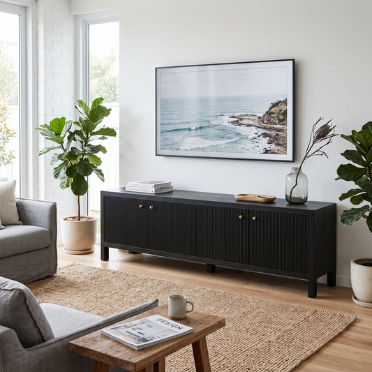 A modern black wooden media console in a bright living room, styled with a framed ocean landscape print on the wall above it, flanked by potted plants and a small wooden side table in the foreground.