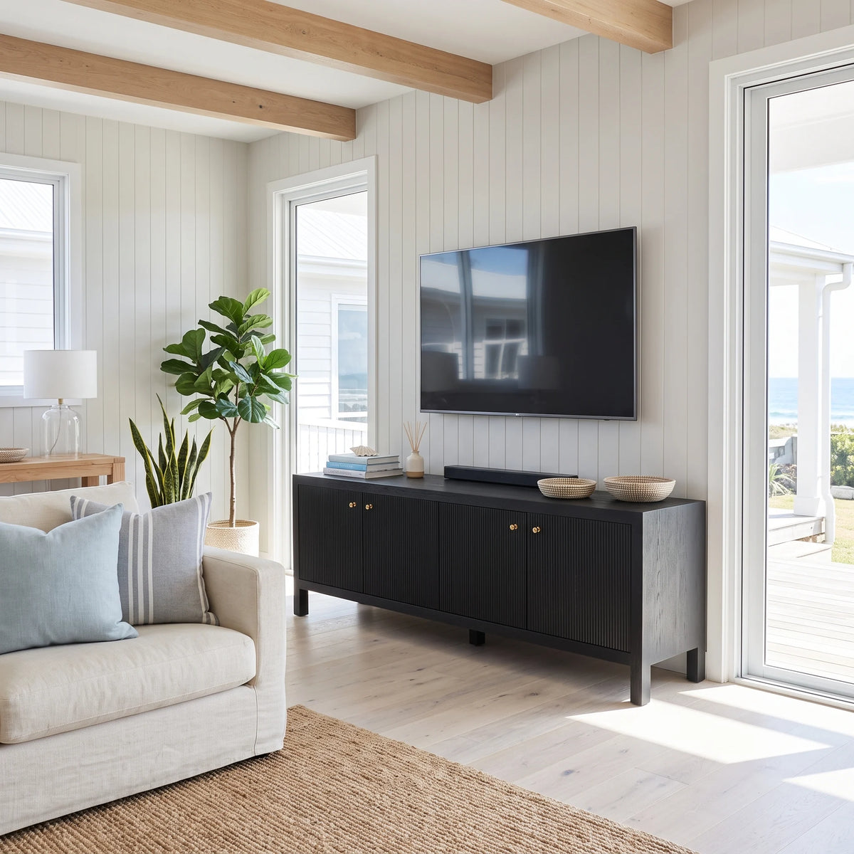 A modern coastal living room featuring a black wooden credenza with brass knobs, a wall-mounted flat-screen TV, and a soundbar. The room has white shiplap walls, light wood ceiling beams, a light beige sofa with blue and gray pillows, and a large green plant.