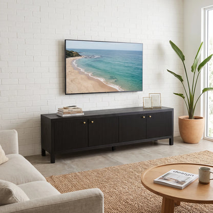 A modern black wood media console with ribbed doors and gold knobs, styled in a bright living room under a wall-mounted TV showing a beach scene.
