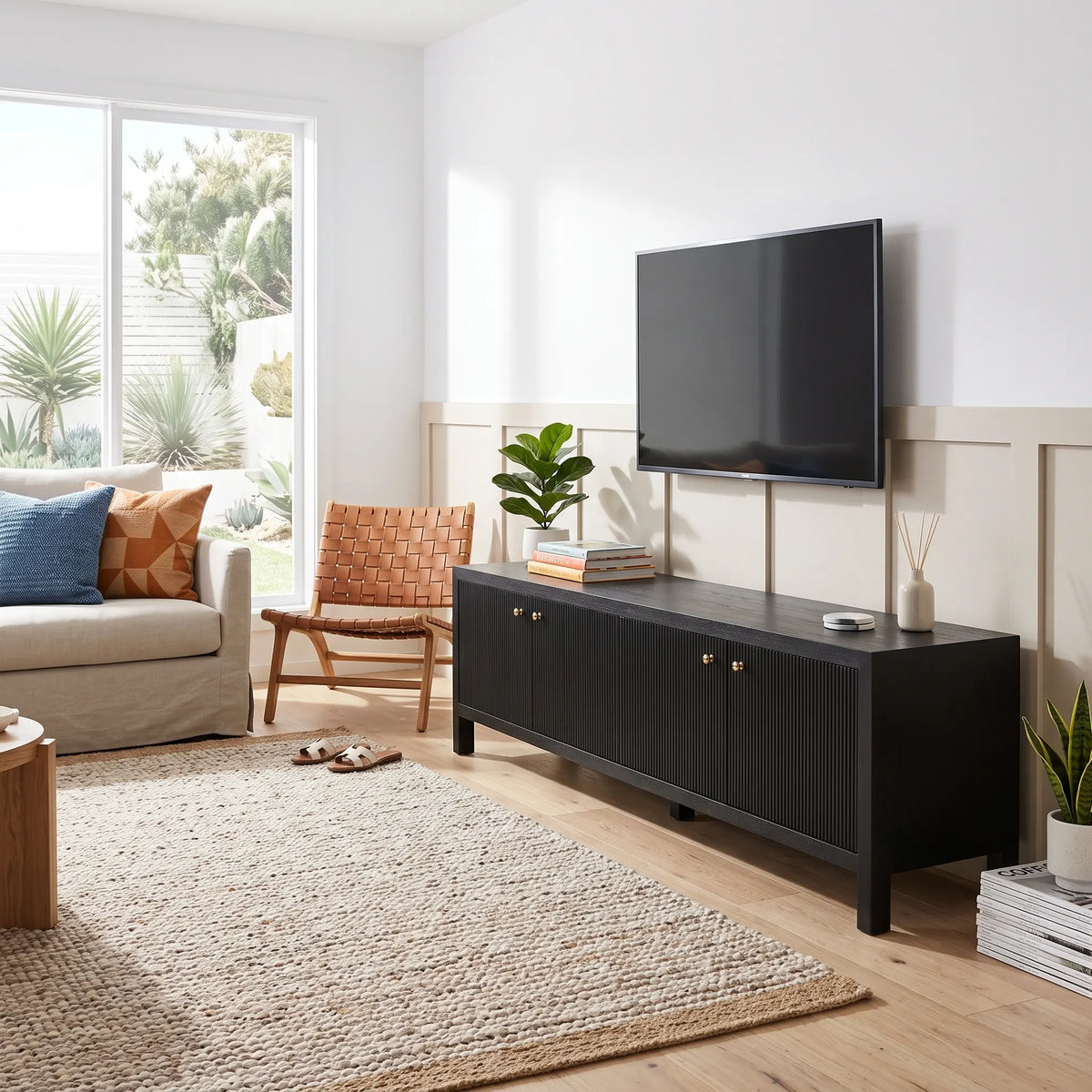 A modern black wood TV stand or media console with two cabinet doors and small gold knobs, shown in a bright living room with a wall-mounted TV, sofa, chair, rug, and plants.