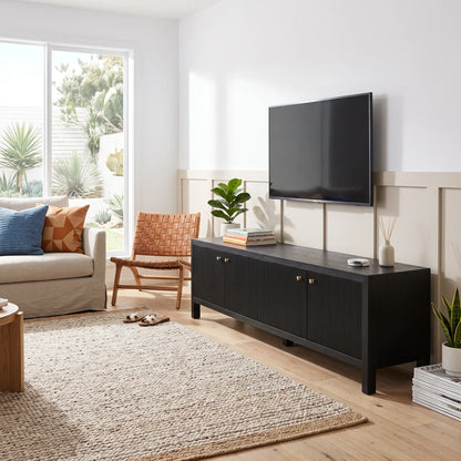 A modern black wood TV stand or media console with two cabinet doors and small gold knobs, shown in a bright living room with a wall-mounted TV, sofa, chair, rug, and plants.