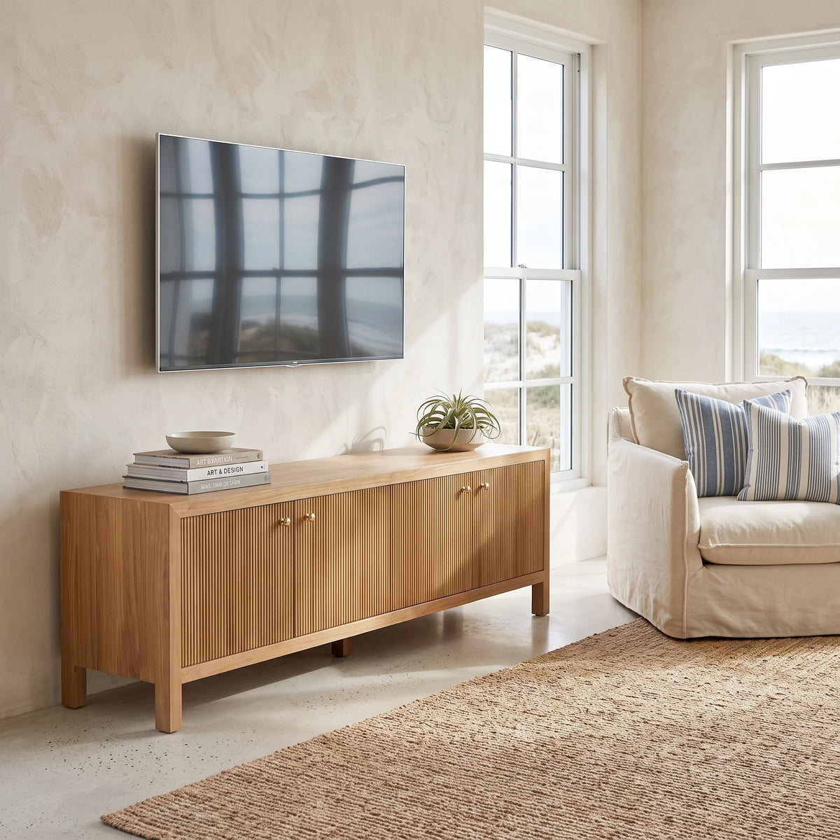 A light natural wood sideboard console with vertical slats and small gold knobs, styled in a bright living room with a wall-mounted TV, books, a plant, and a cream armchair.