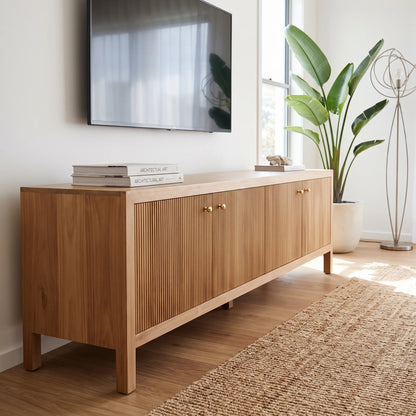 A light oak wood sideboard cabinet with vertical fluted doors and small gold knobs, styled in a bright living room next to a window with a potted plant and a TV mounted on the wall.