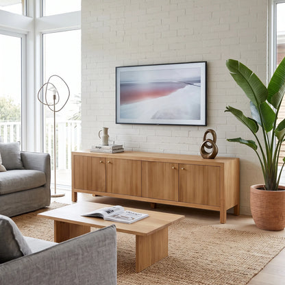 A mid-century modern wooden sideboard cabinet in a bright, contemporary living room. The light oak cabinet features vertical slatted doors and small round brass knobs, styled with a framed print, a ceramic vase, and a sculptural object.