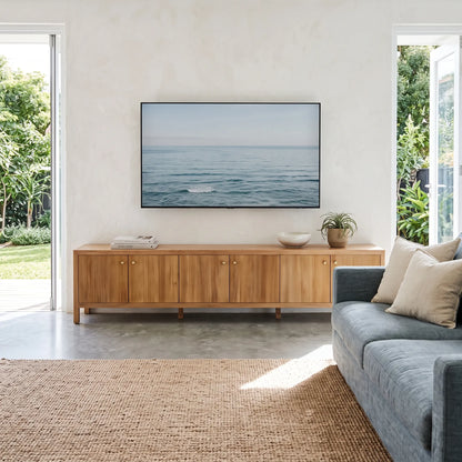 A framed horizontal ocean landscape print hangs on a textured off-white wall above a light wood sideboard in a modern living room with a blue-gray sofa and woven rug.