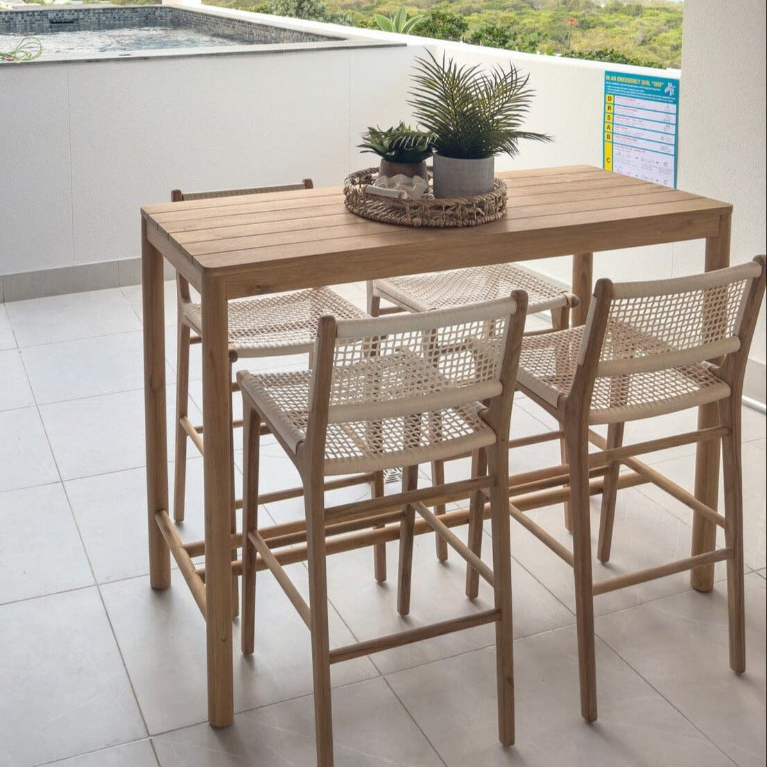 A teak outdoor bar table with a slatted top design, accompanied by high stools, set on a tiled floor with a scenic view in the background.