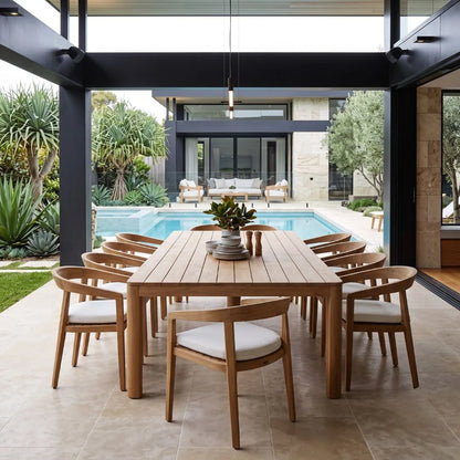Outdoor dining area with wooden table and chairs on a patio, pool, and garden in the background.