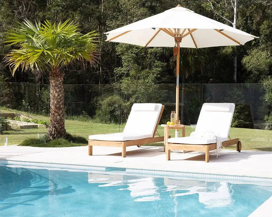 Two white lounge chairs under a large umbrella by a pool with a palm tree in the background.