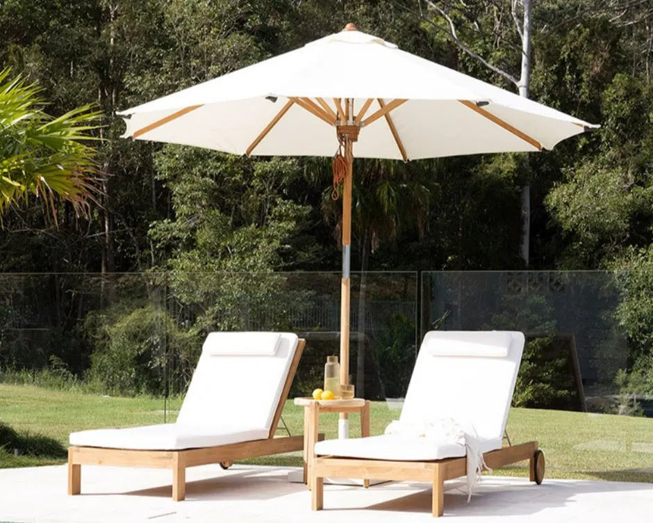 Two wooden lounge chairs with white cushions under a large outdoor umbrella in a garden setting.