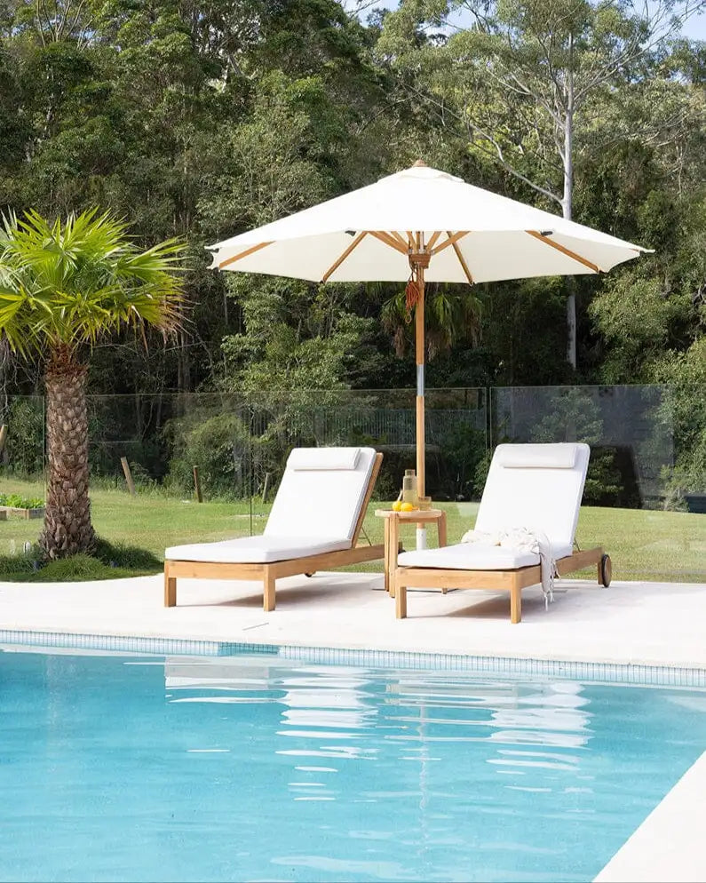 Two white lounge chairs with a beige umbrella by a poolside with greenery in the background.