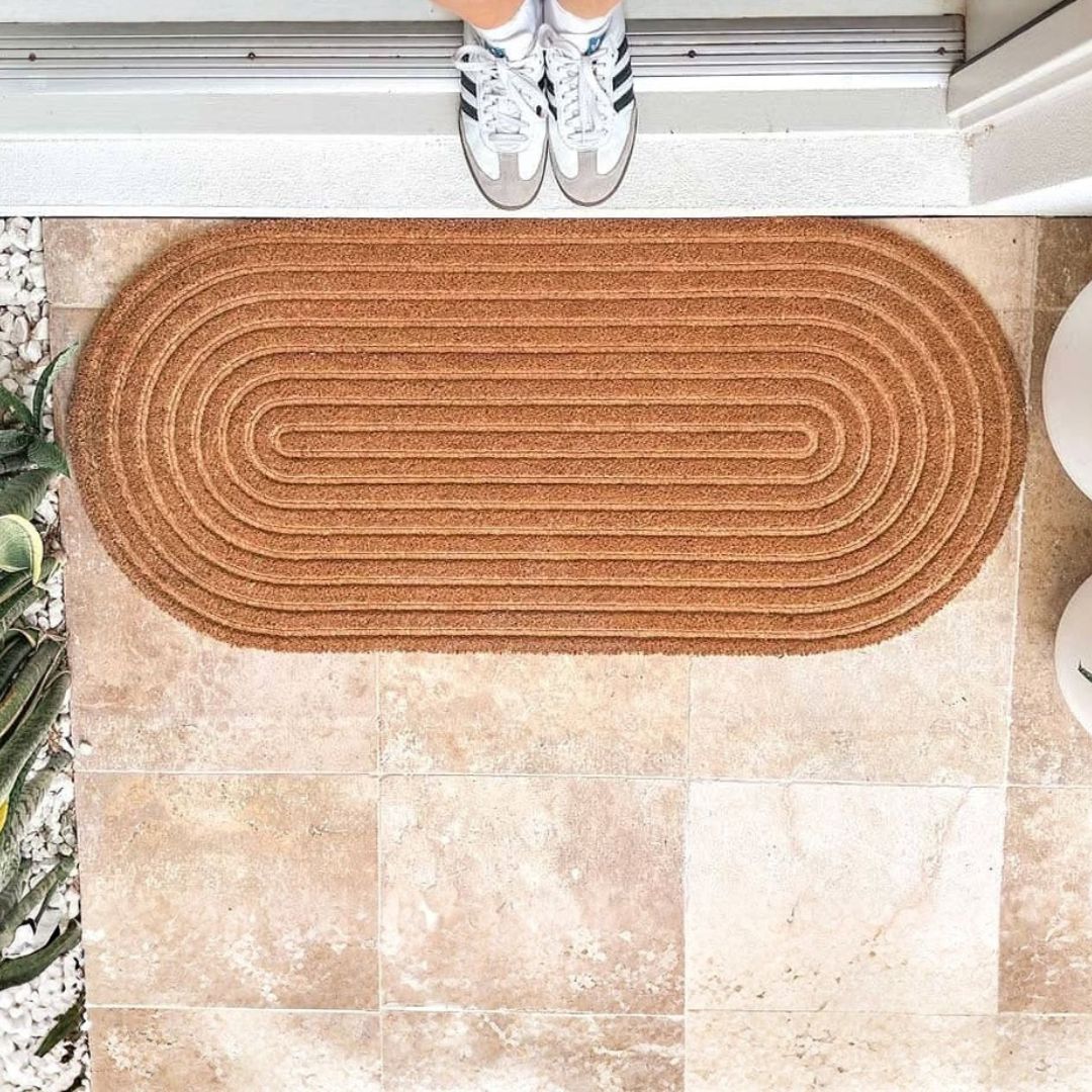 Brown doormat on a tiled floor with a person wearing white sneakers standing on it.