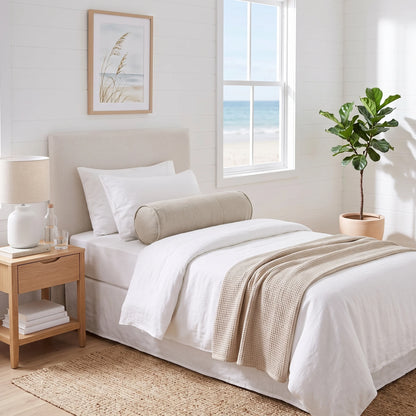 A neatly made bed in a bright, modern bedroom with white linens, a beige cylindrical bolster pillow, a beige textured throw blanket, a light wood nightstand with a white lamp, and a potted fiddle-leaf fig near a window overlooking the ocean.