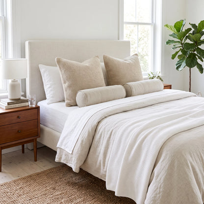 A neatly made bed in a bright bedroom, dressed in off-white and beige bedding with matching pillows, including two large textured pillows and a cylindrical bolster pillow. A wooden nightstand with a white lamp sits beside the bed, and a green plant is visible near the window.