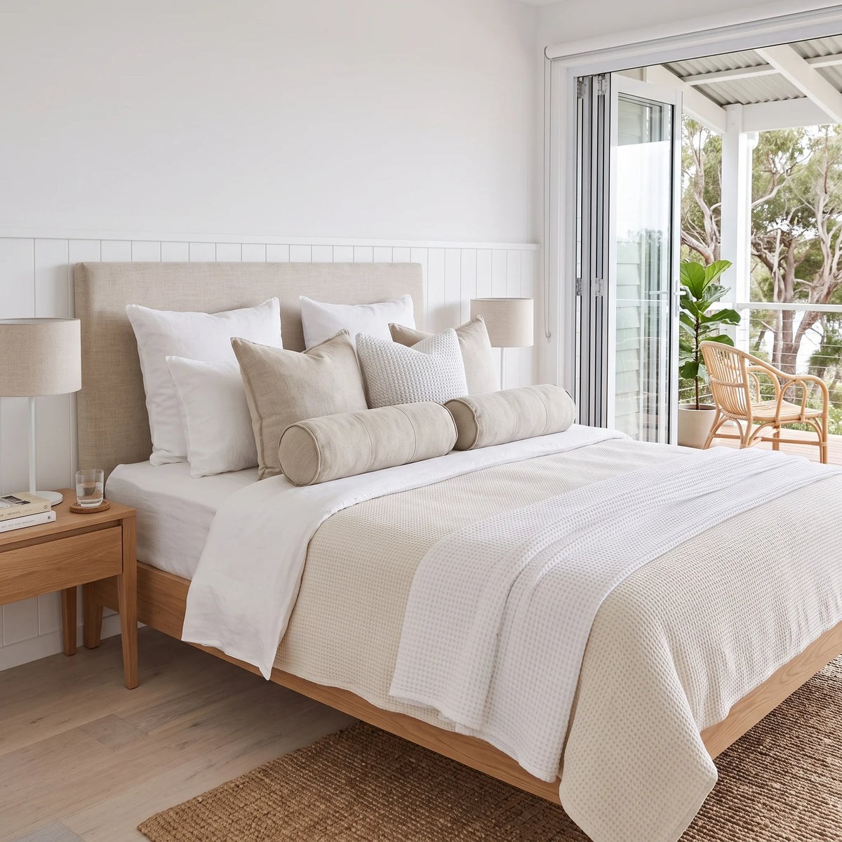 A neatly made bed in a bright, modern bedroom featuring a light beige upholstered headboard, white and cream bedding, matching pillows, a wooden nightstand with a lamp, and sliding glass doors leading to a balcony.