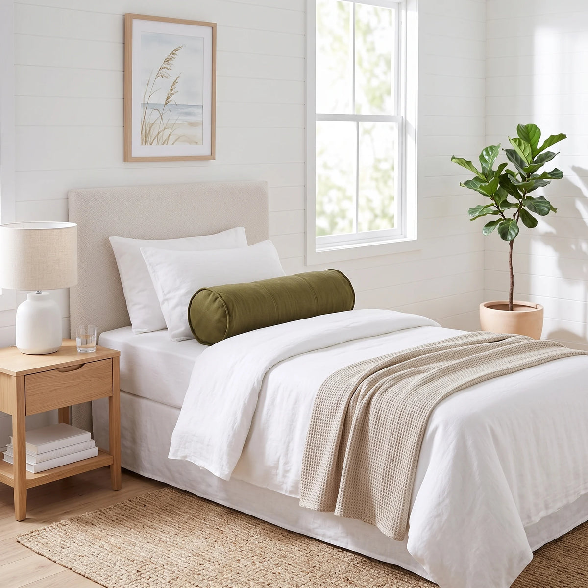 A styled bedroom scene showing a bed with white linens, a beige textured throw blanket, and an olive green cylindrical bolster pillow. The bed is set in a bright, modern bedroom with a light wood nightstand, a white table lamp, a potted fiddle-leaf fig plant, and a framed print on the wall.