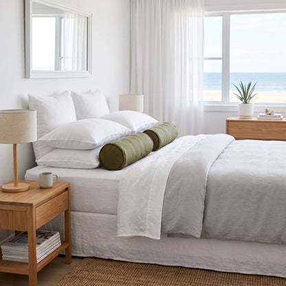 A neatly made bed in a bright, minimalist beach-style bedroom. The bedding is predominantly white with a cylindrical olive green bolster pillow in the center.