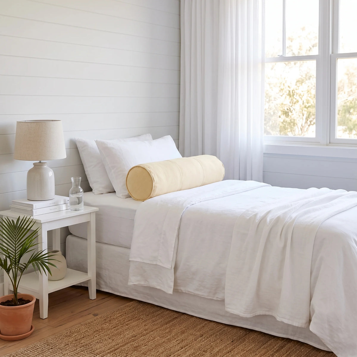 A neatly made bed with white linens and two cylindrical pillows, including one pale yellow bolster pillow, in a bright bedroom with white shiplap walls, sheer curtains, a small white nightstand, a lamp, and a potted plant.