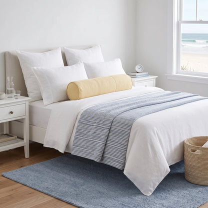 A neatly made bed in a bright bedroom with white bedding, a light blue striped throw blanket, and a round yellow bolster pillow. A window looks out to the ocean, and a blue rug covers the wood floor.