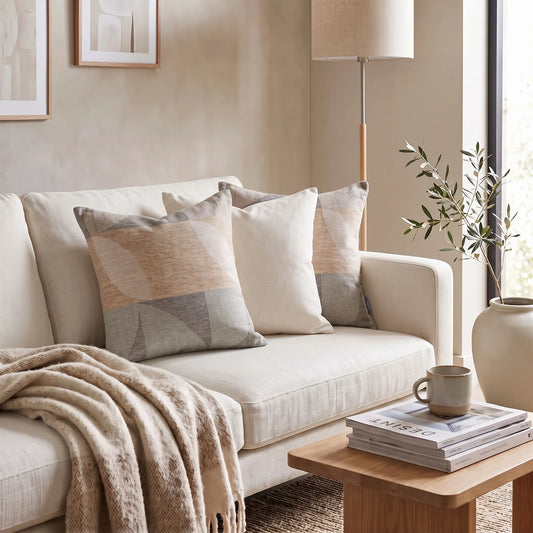 A styled living room scene with a cream sofa, several throw pillows in beige, cream, and gray geometric patterns, a beige throw blanket, a small wooden side table with a mug and books, a floor lamp, and a vase with branches.