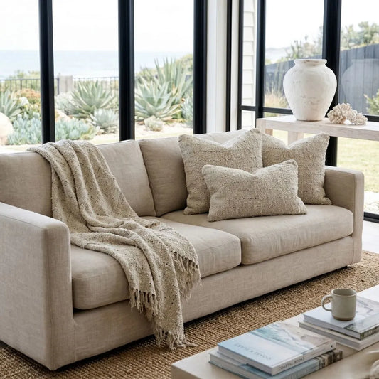 Beige sofa with pillows and a blanket in a living room with large windows.