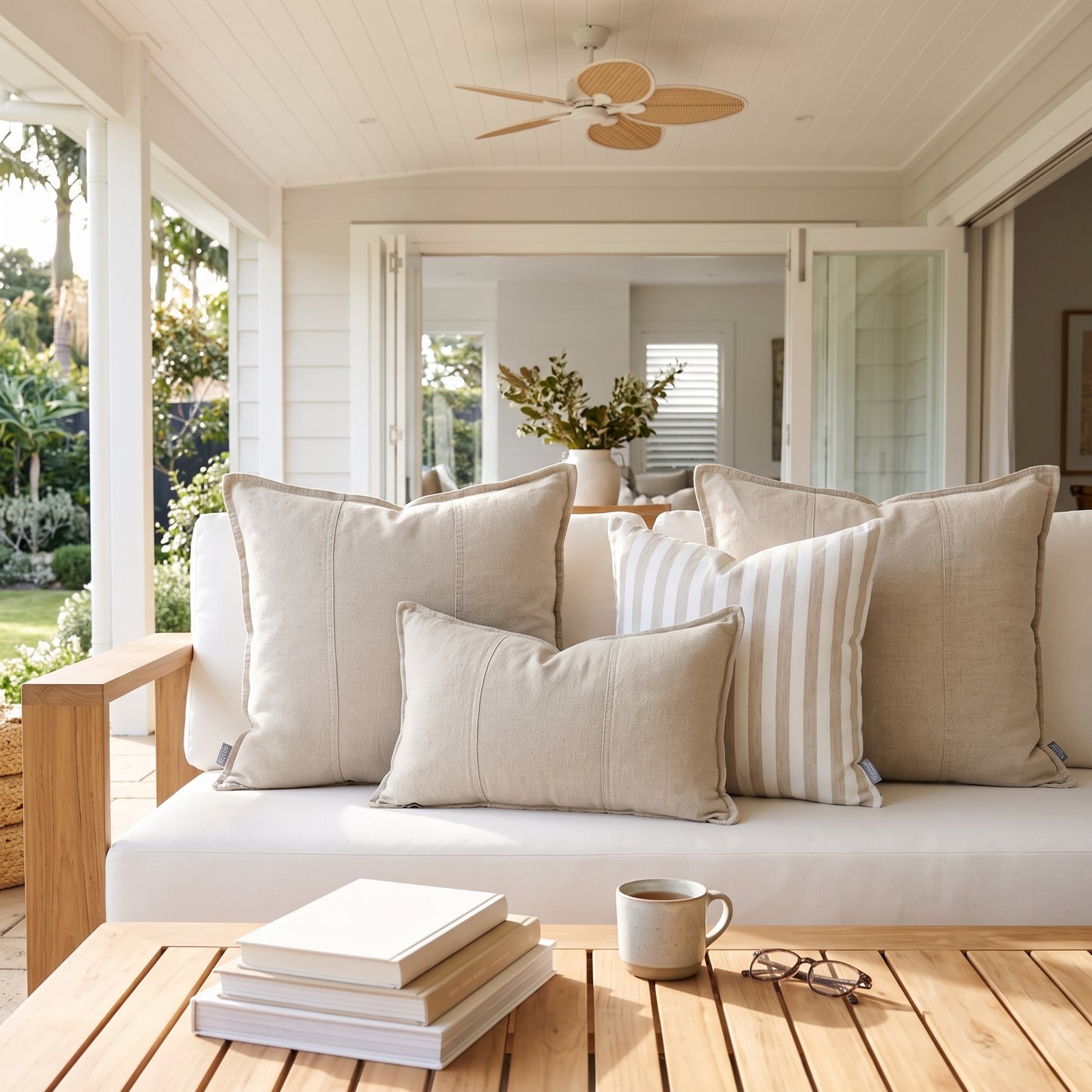 A set of neutral-toned throw pillows, including solid beige and cream-and-white striped pillows, arranged on a white outdoor sofa on a covered porch with a wooden slat coffee table in the foreground.